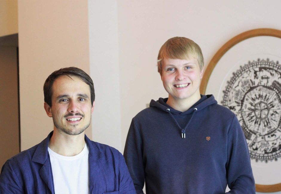 Loris und Joel befinden sich im Eingangsbereich von Ergon. Hinter ihnen ist ein Bild zu sehen, das aus einem runden Scherenschnitt besteht. Der Scherenschnitt bildet die Geschichte von Ergon ab. Loris und Joel befinden sich im Eingangsbereich von Ergon. Hinter ihnen ist ein Bild zu sehen, das aus einem runden Scherenschnitt besteht. Der Scherenschnitt bildet die Geschichte von Ergon ab.