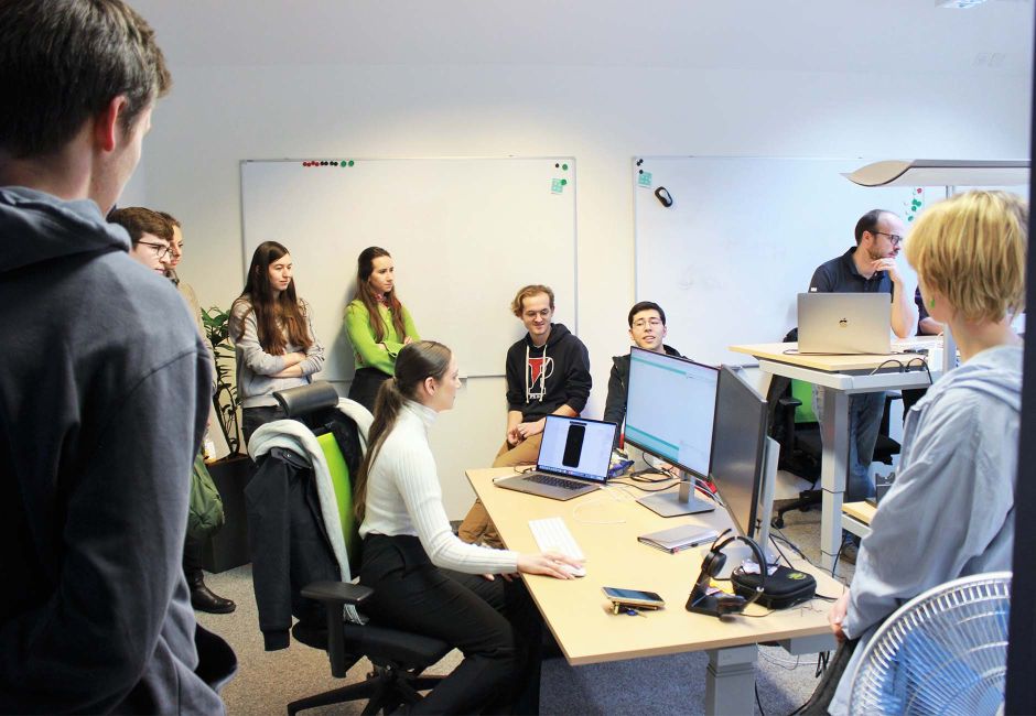 Neun Praktikant:innen haben sich in einem Büro versammelt und stehen rund um einen Arbeitstisch herum. Am Tisch sitzt eine Praktikantin und erklärt etwas auf ihrem Bildschirm. Rechts im Bild ist ein Software-Entwickler zu sehen, der am Stehtisch arbeitet. Neun Praktikant:innen haben sich in einem Büro versammelt und stehen rund um einen Arbeitstisch herum. Am Tisch sitzt eine Praktikantin und erklärt etwas auf ihrem Bildschirm. Rechts im Bild ist ein Software-Entwickler zu sehen, der am Stehtisch arbeitet.